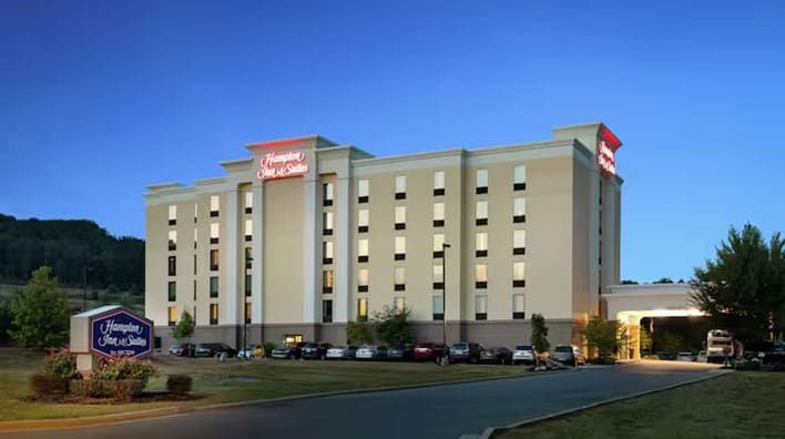 Multi-story Hampton Inn & Suites Adairsville hotel building with exterior lighting, a parking lot filled with cars, and trees surrounding the property at dusk.