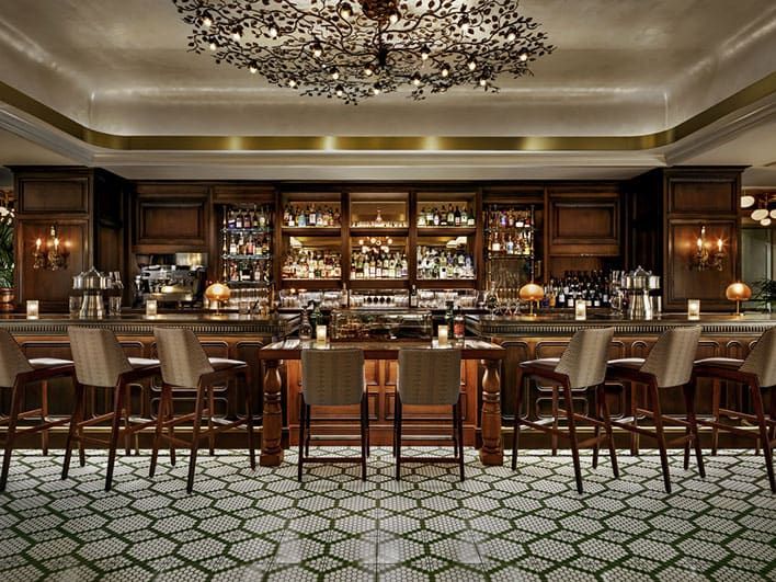 A polished bar interior with wooden stools, a patterned tile floor, shelves filled with bottles, and warm lighting under an ornate ceiling fixture.