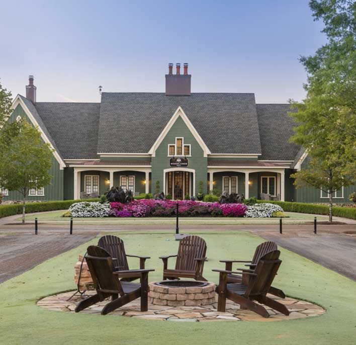 Large green house with white trim, a flower garden in front, and a circular fire pit area with four wooden chairs on a lawn in the foreground.