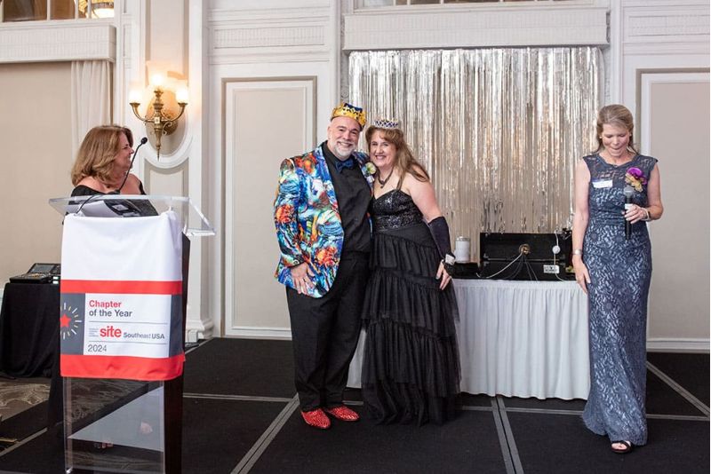 Four adults stand on a stage at an event; one man wears a crown and colorful jacket, while others wear formal dresses. A podium with a “Chapter of the Year 2024” sign is visible.