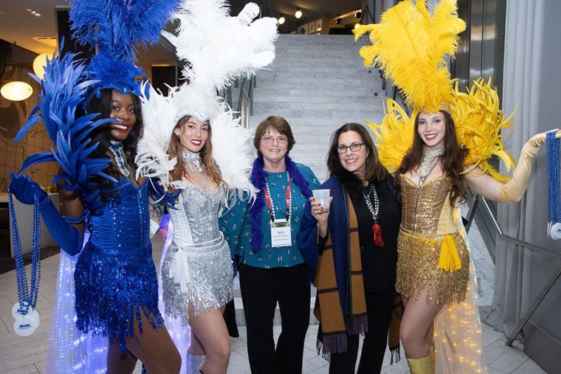 Two women stand between three showgirls in colorful feathered costumes on a staircase, posing and smiling for the camera at an indoor event.