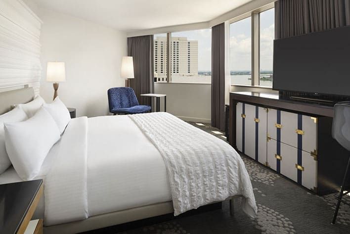 Corner Suite room at Le Méridien New Orleans with a large bed, blue chair, desk, dresser, flat-screen TV, and floor-to-ceiling windows overlooking city buildings.