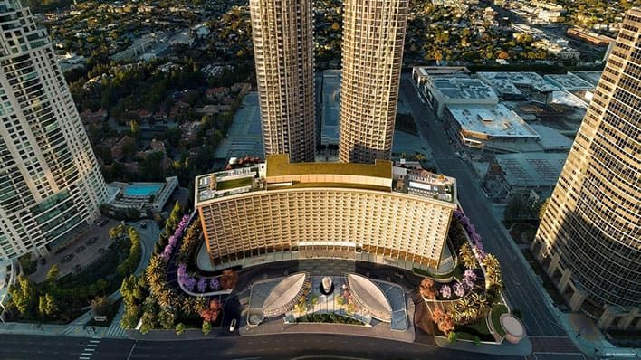 Aerial view of the Fairmont Century Plaza high-rise hotel with two tall towers behind it, surrounded by city streets, buildings, and trees.
