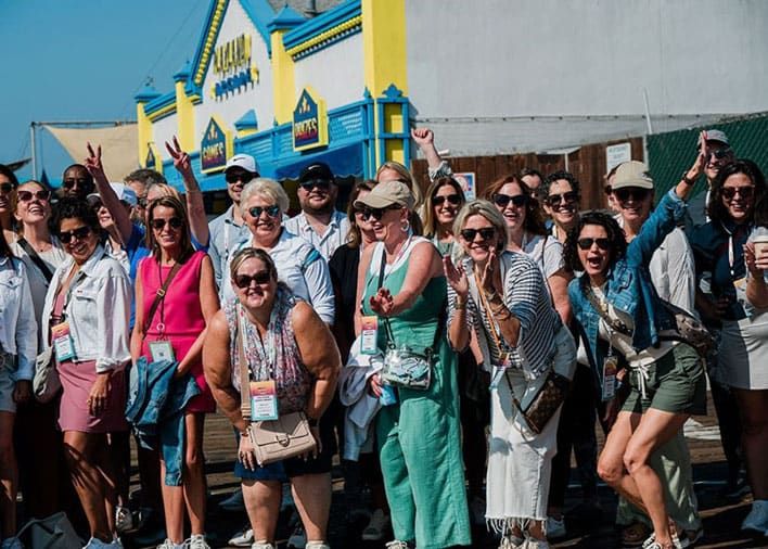 A large group of adults pose and smile for a photo outdoors in front of a colorful building on a sunny day.