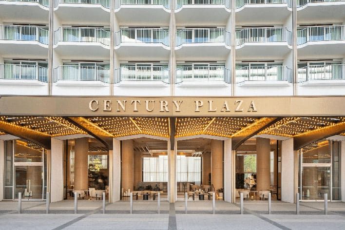 The entrance of Fairmont Century Plaza hotel features a modern façade with a gold canopy, illuminated lights, and rows of balconies above.