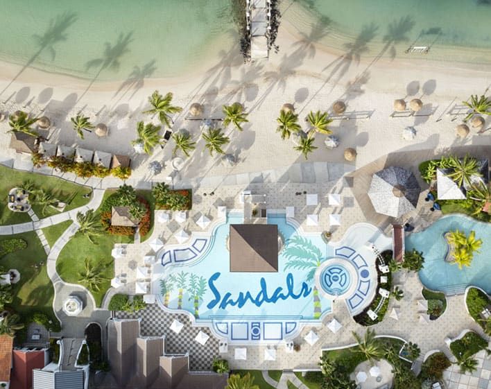 Aerial view of a beachfront resort with a pool featuring the word "Sandals," surrounded by palm trees, lounge chairs, and a sandy beach next to clear turquoise water.