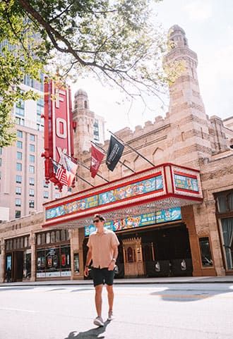 Person walking past the historic Fox Theater with intricate architecture and colorful marquee signs, located in an urban setting with tall buildings and trees.