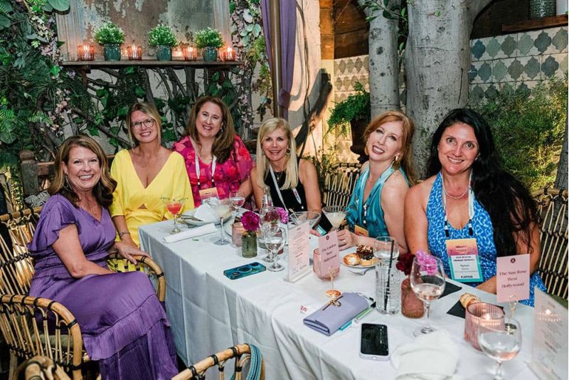 Six women sit together at a decorated table outdoors, smiling at the camera, with drinks and food in front of them.