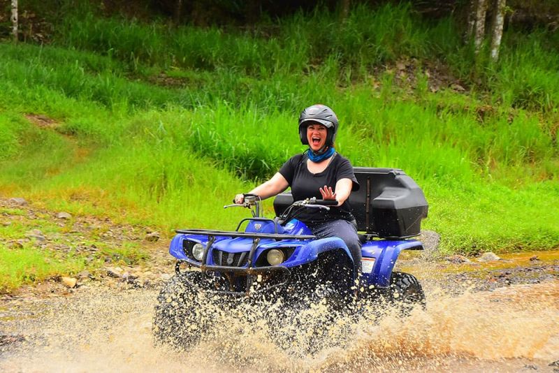 Woman in a helmet riding an ATV through a creek, with water splashing around, surrounded by greenery.