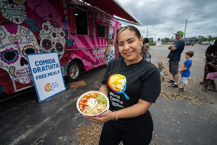 A woman holding a bowl of food stands in front of a World Central Kitchen food truck with a sign offering free meals. There are several other people in the background, including children, standing in line.