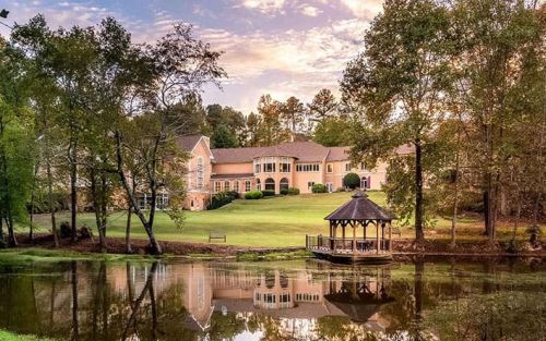 Large resort with cream facade and multiple windows, surrounded by trees with a small gazebo by a reflecting pond in the foreground.