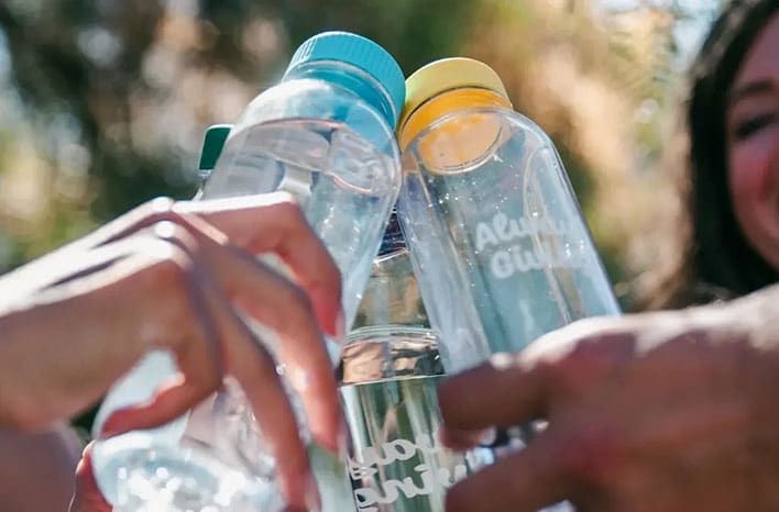 Several hands hold four clear, reusable Fill it Forward water bottles with different colored caps in an outdoor setting.