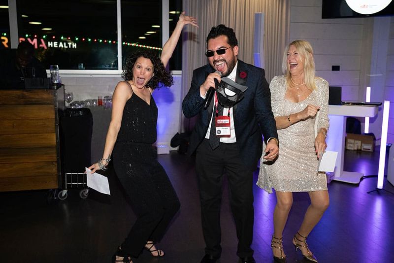 Three people are having fun, with one man singing into a microphone and two women dancing beside him at a party.